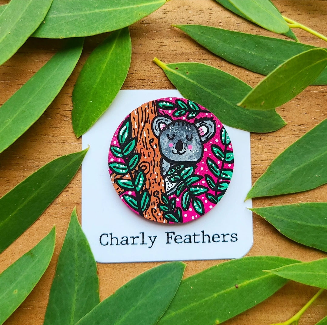 Circular needle minder with a koala design on a card labeled 'Charly Feathers', surrounded by green leaves on a wooden surface.