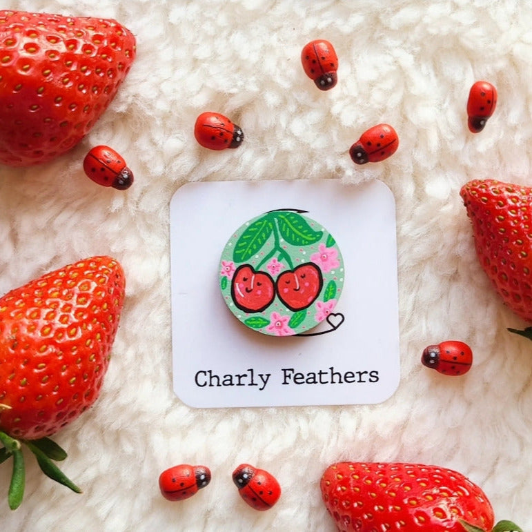 needle minder with cherry design on a card labeled 'Charly Feathers' surrounded by strawberries and ladybugs on a fluffy white surface.