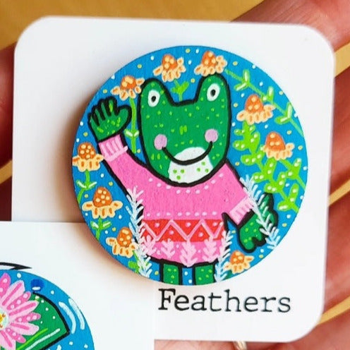 Colorful illustration of a frog wearing a sweater with flowers around it, held by a hand.