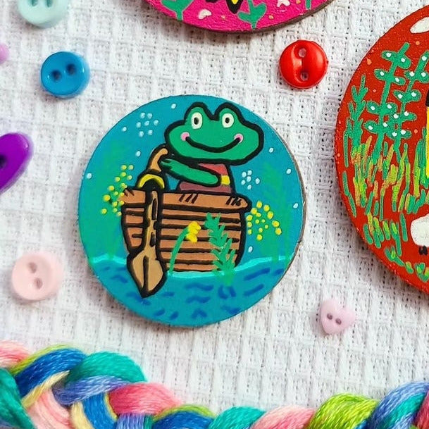 illustration of frog in a boat on a small needle minder surrounded by floss and buttons