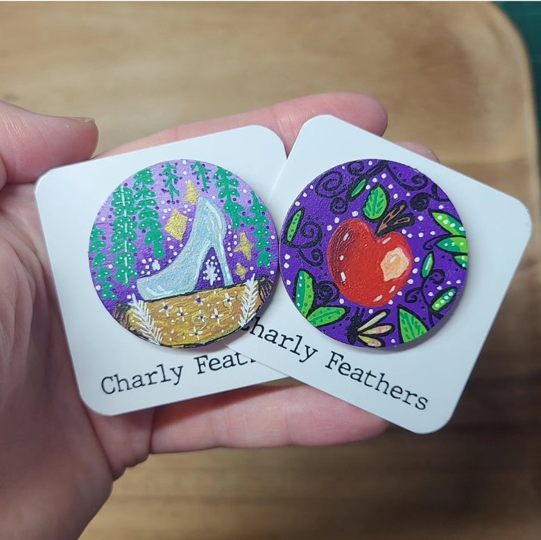 Two round needle minders with colorful designs held by a hand on a wooden surface.