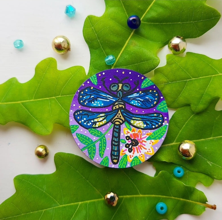 Colourful dragonfly-themed needle minder on green leaves with decorative beads.