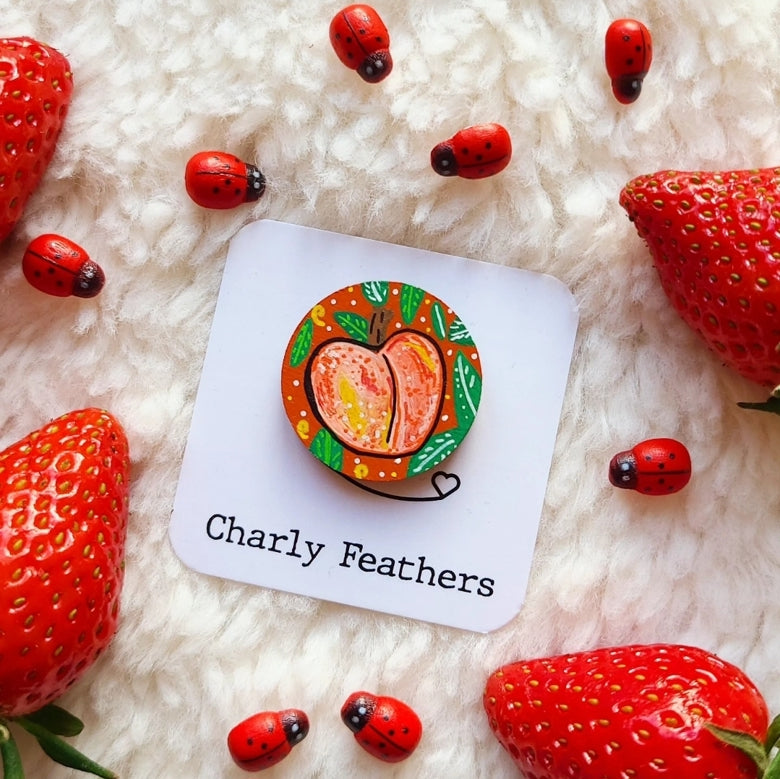 Decorative needle minder with a peach design on a white card, surrounded by strawberries and ladybugs on a fluffy white surface.