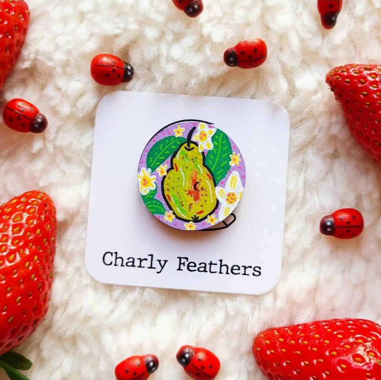 Colorful needle minder with a pear design on a white card labeled 'Charly Feathers', surrounded by strawberries and ladybugs on a fluffy white background.