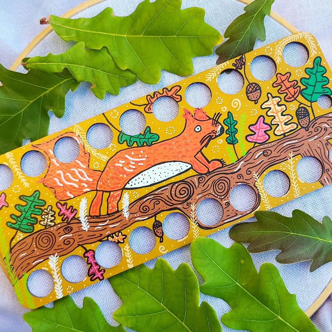 Colorful wooden thread organiser with a squirrel design on a blue background with green leaves.