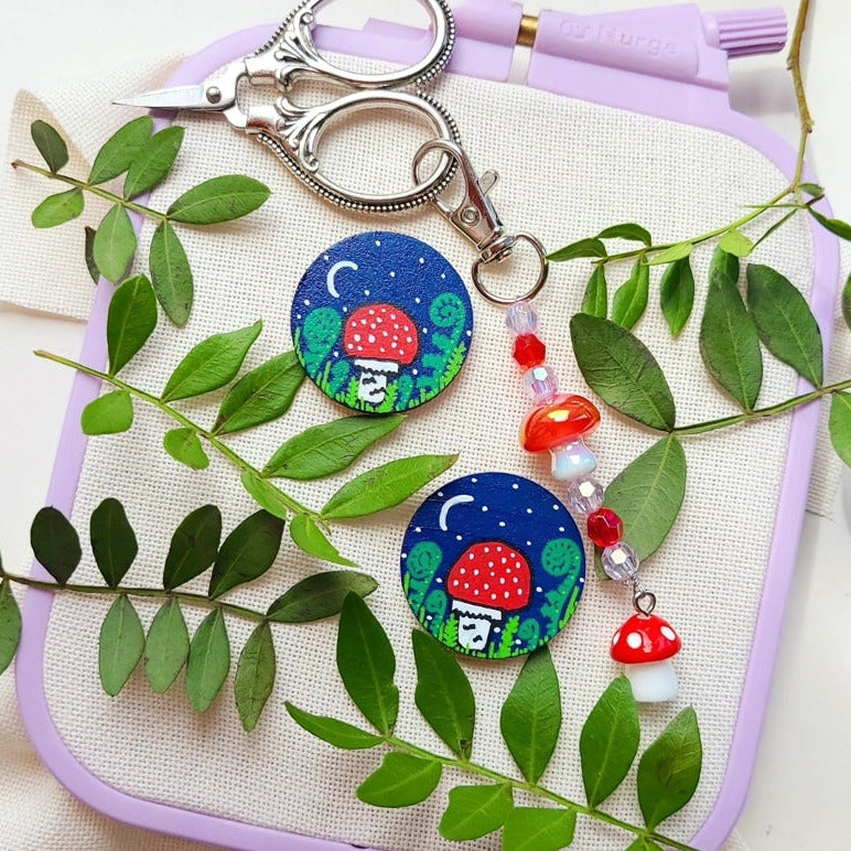 Two colorful mushroom-themed needle minders on fabric with green leaves.