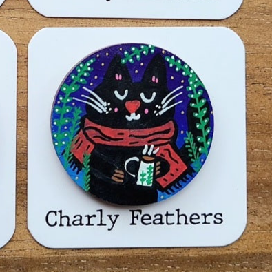 Circular badge with a cat design holding a mug, branded 'Charly Feathers', on a wooden surface.